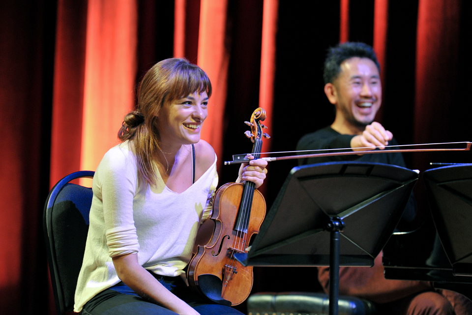 A female student, holding a violinist, sitting next to another student, who are both laughing.